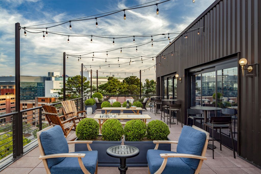 Graduate by Hilton Charlottesville, outdoor seating, chess, wooden lounge seating, blue cushions, potted plants, bistro string lights, black bistro table, gold outdoor wall mount lighting, red plaid couch cushions, black bar table, black bar stools, wooden outdoor tables