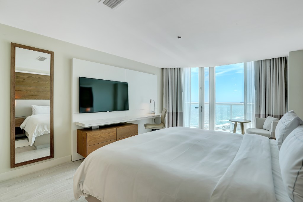 Beach House Fort Lauderdale, a Hilton Resort - Oceanfront Two Bedroom Suite - Bedroom, large bed, TV, mirror, chair and table, long desk surface under tv long the wall, balcony overlooking the ocean