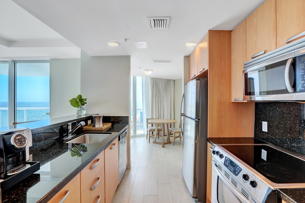 Beach House Fort Lauderdale, a Hilton Resort - Oceanfront Two Bedroom Suite - Kitchen, countertop with sink, coffemake and cabinets with dishwasher and on the opposite side cabinets with refrigerator, stove and microwave - table and chair in the distance