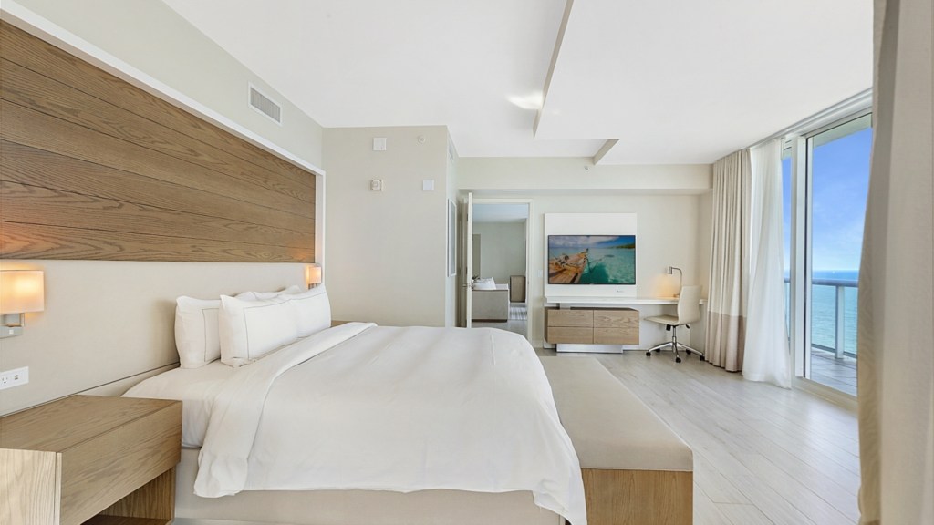 Beach House Fort Lauderdale, a Hilton Resort - Penthouse Bedroom, bed with side tables and lamps overlooking ocean view, tv and desk with desk chair in the corner door to living room