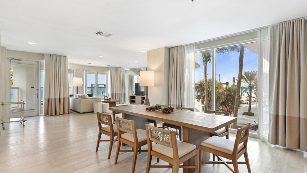 Beach House Fort Lauderdale, a Hilton Resort - Presidential Suite Living Room, long wooden dining table and chairs, couch, TV and lamps in the background, view of glass doors to outside and to the beach