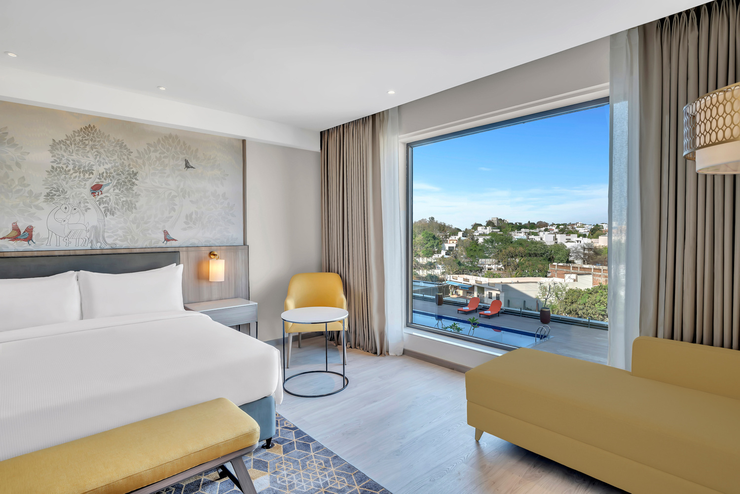 Suite with wall art above the bed's headboard, bed with white bedding with a yellow bench at the foot of the bed and a side table with wall sconce light, Chaise lounge couch seating area with a window view, sitting area with a yellow chair and round white table next to the window