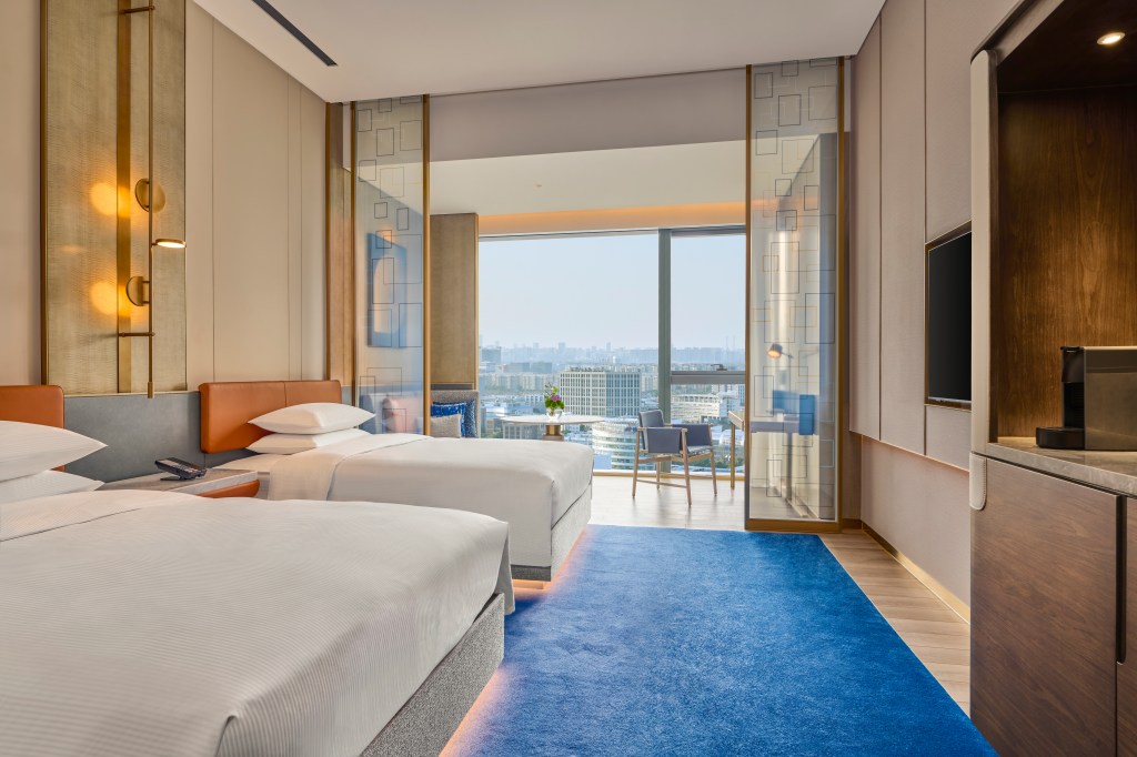 two queen beds with side tabels and lamps, blue carpet and balcony with seating and a table overlooking city