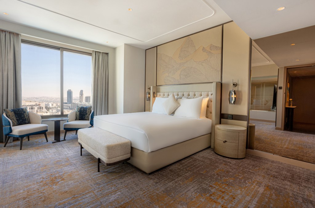 Queen bed with side table and table and chairs near window overlooking city