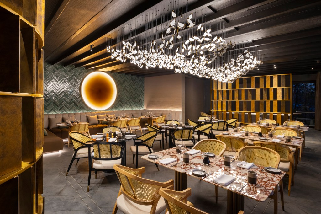 dining area with yellow and black chairs around tables with place settings in the back of the room there is a large booth area with 3 tables and a room partian off to the side decorative light fixture hangs from the ceiling