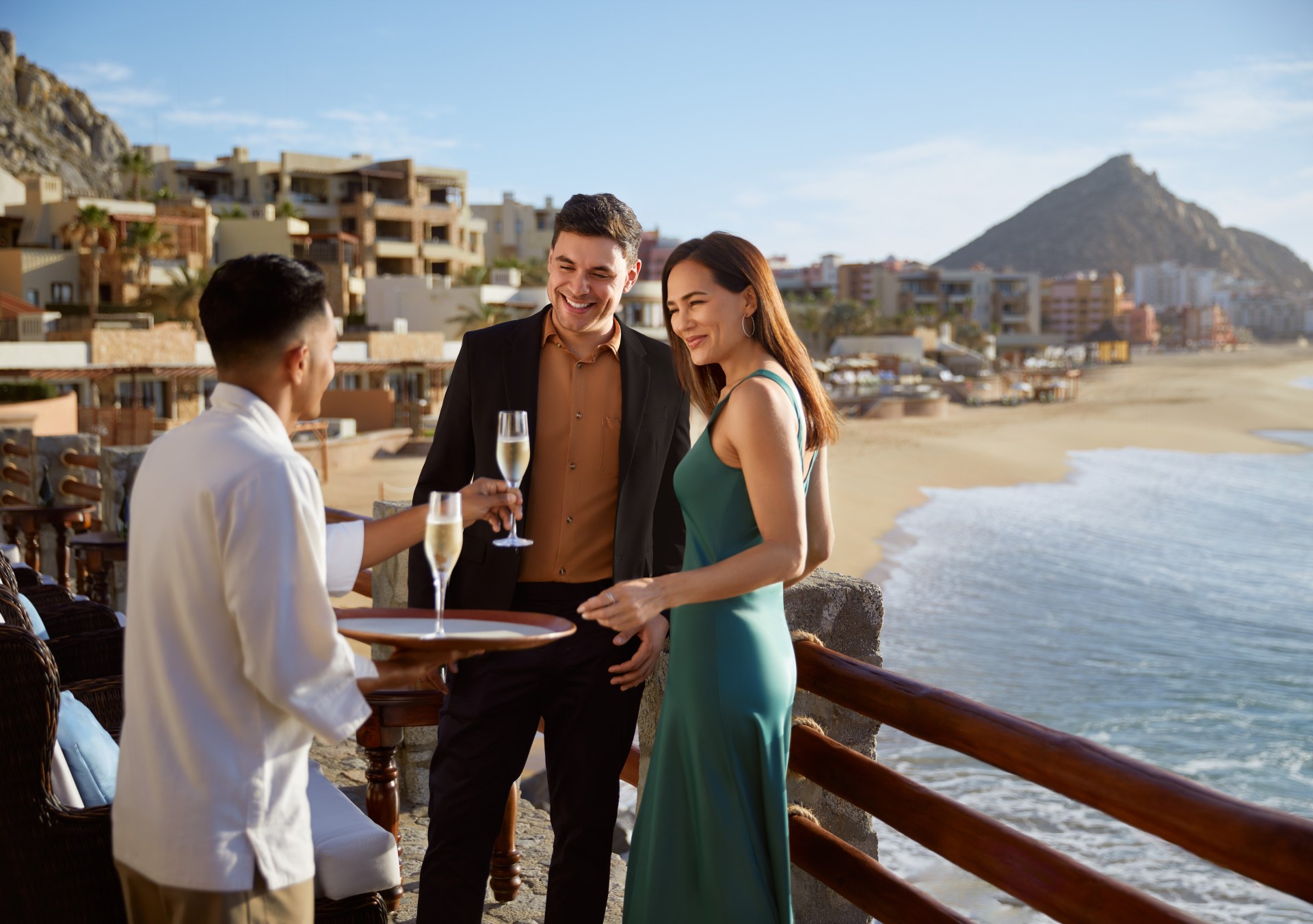 Waldorf Astoria Los Cabos Pedregal - couple being served sparkling wine by a waiter on a balcony overlooking ocean, hotel and mountain the background