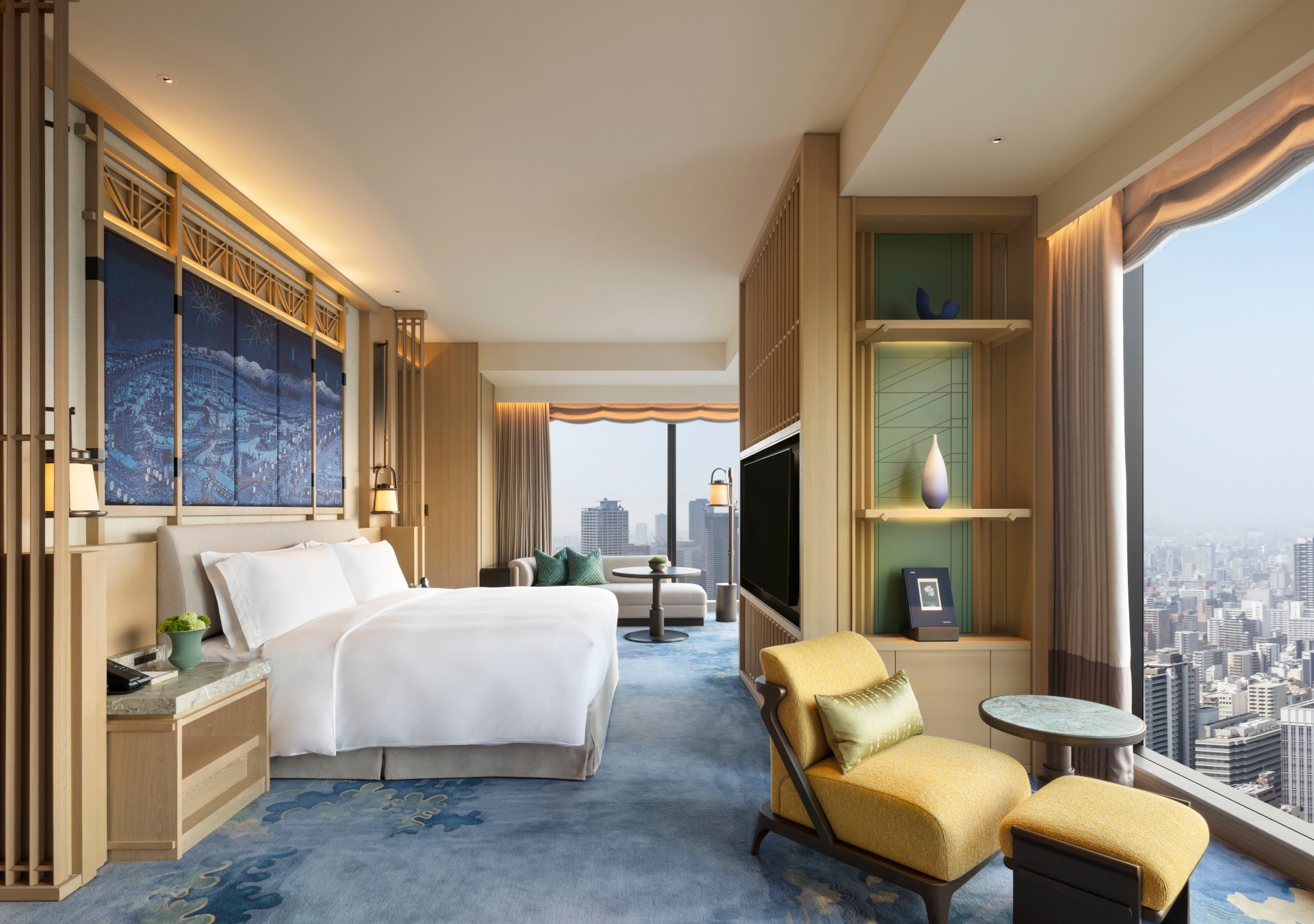 Waldorf Astoria Osaka - Presidential Suite Bedroom, large windows overlooking city skyline, bed with white bedding, large blue city artwork hanging on the wall over bed, table and chair seating area with yellow chair, cream sofa