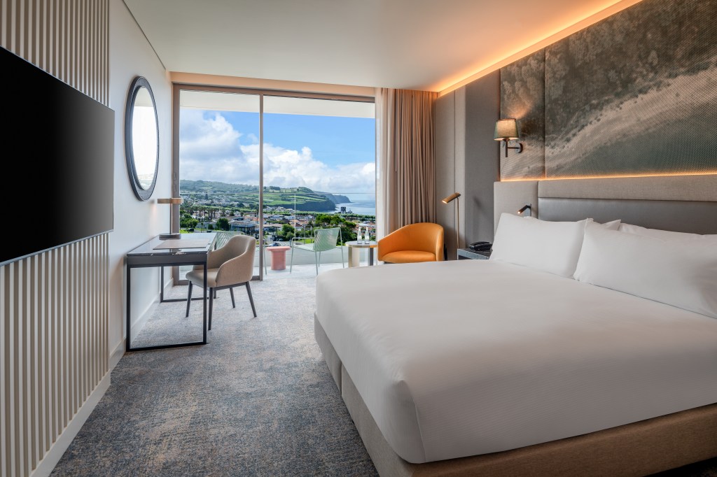 DoubleTree by Hilton Lagoa Azores, King Guest Room with view of the coast and an outdoor balcony with seating, orange chair, wall art, desk with seat, round mirror hanging on wall, mounted television on slotted textured wall