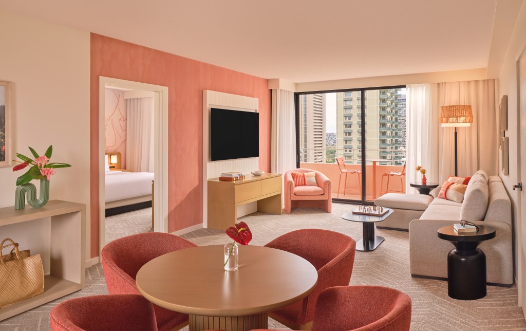 The Ambassador Hotel of Waikiki, Tapestry Collection by Hilton - Suite Living Room - Credit - Michael Clifford, pink and beige living room with couch, table and chairs, coffee table with chess board, view of balcony an view of bedroom area