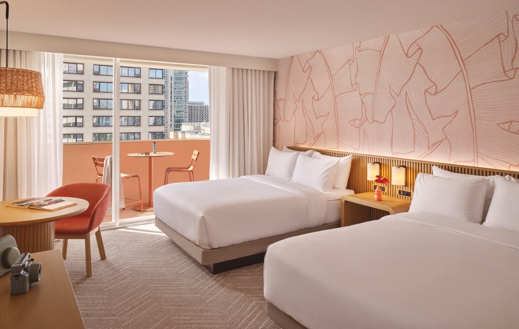 The Ambassador Hotel of Waikiki, Tapestry Collection by Hilton - Two Queen Beds Guest Room - Credit - Michael Clifford, bedroom with double beds with nightstand and sconces in the center, table and chair and view of balcony with outdoor table and chaiars.
