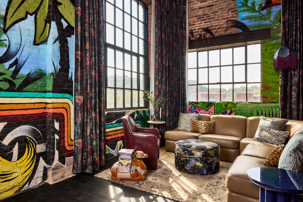 Lobby with painted brick wall and leather chair and l-shaped couch with throw pillows next to an ottoman and end tables all sitting on top of a patterned rug