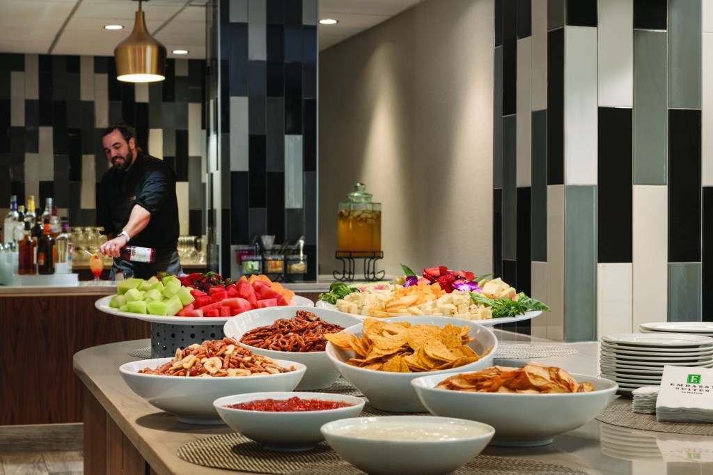 Snack buffet table with fruit and chips with salsa and dip along with pretzels and cheese next to a drink bar