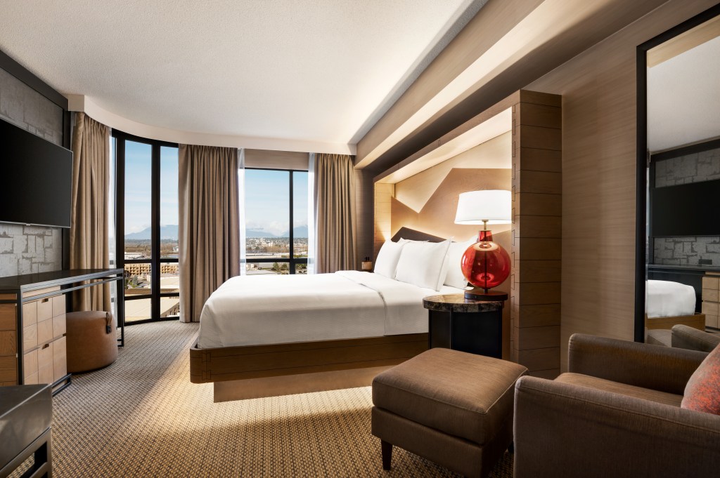 Hilton Vancouver Airport 1 King Bed, King be with white bedding in a room with a red table lamp on a round end table with a lounge chair and television windows looking out to the city with mountains in the distance