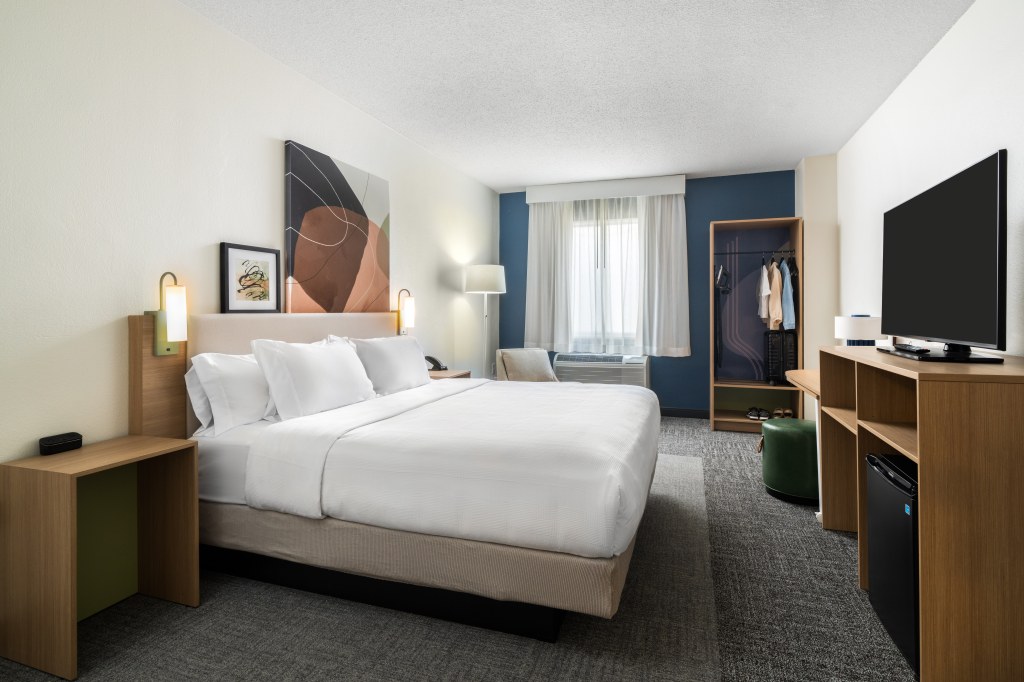 Spark by Hilton Chattanooga Downtown King Bed with artwork hanging on the wall above and mounted sconce lights next to a night stand on each side of the bed and a lounge seat next to a window and an armoire closet to hang clothing facing a desk and media center with a seat and mini refridgerator