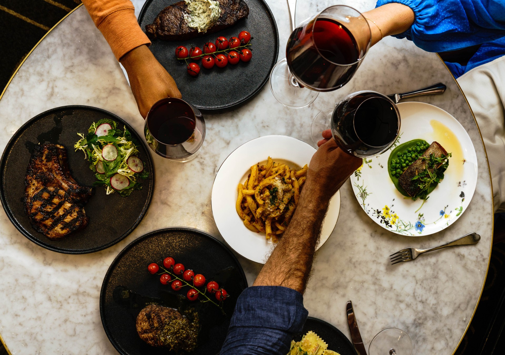 Waldorf Astoria Chicago Dishes from Brass Tack, table with plated dishes and glasses of red wine, tomatoes on vine, grilled pork, ravioli, people toasting with red wine in stemmed glassware