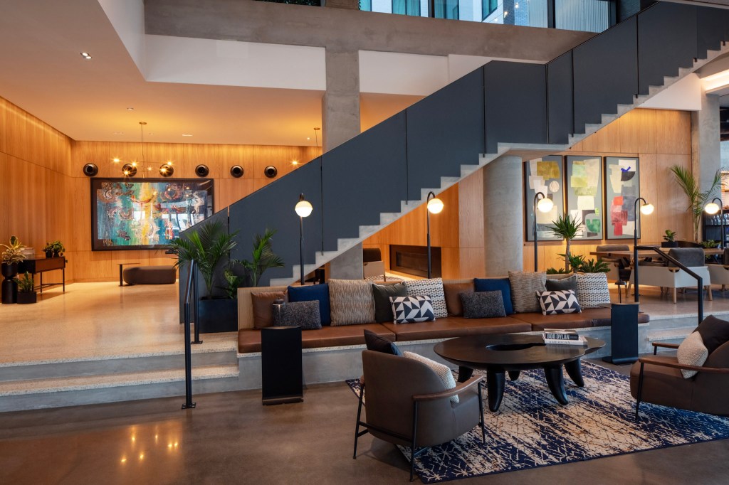Canopy by Hilton Nashville Downtown The Gulch Lobby, sitting area with floor lamps next to a long cushioned bench and chairs with tables with lamps and books, hanging artwork,