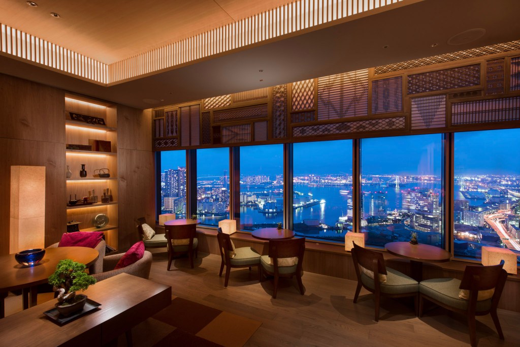 Elegant executive lounge at Conrad Tokyo with wood paneling, artful shelving, and panoramic night views of Tokyo Bay through floor-to-ceiling windows. Executive Lounge Bay View, Conrad Tokyo. Hilton