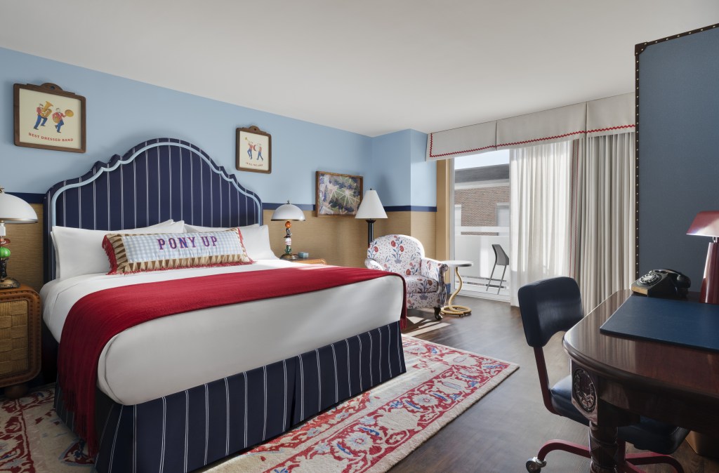 Graduate by Hilton Dallas Graduate, Guest Room with balcony and king bed with framed artwork hanging above the blue striped headboard with white bedding and a red throw blanket and end tables with a lamp on top next to a balcony door and a desk with chair