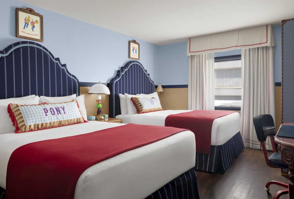 Graduate by Hilton Dallas, Guest Room with two beds with framed artwork hanging above their blue striped headboards with white bedding and a red throw blanket and an end table with a lamp on top next to a window and a desk with chair