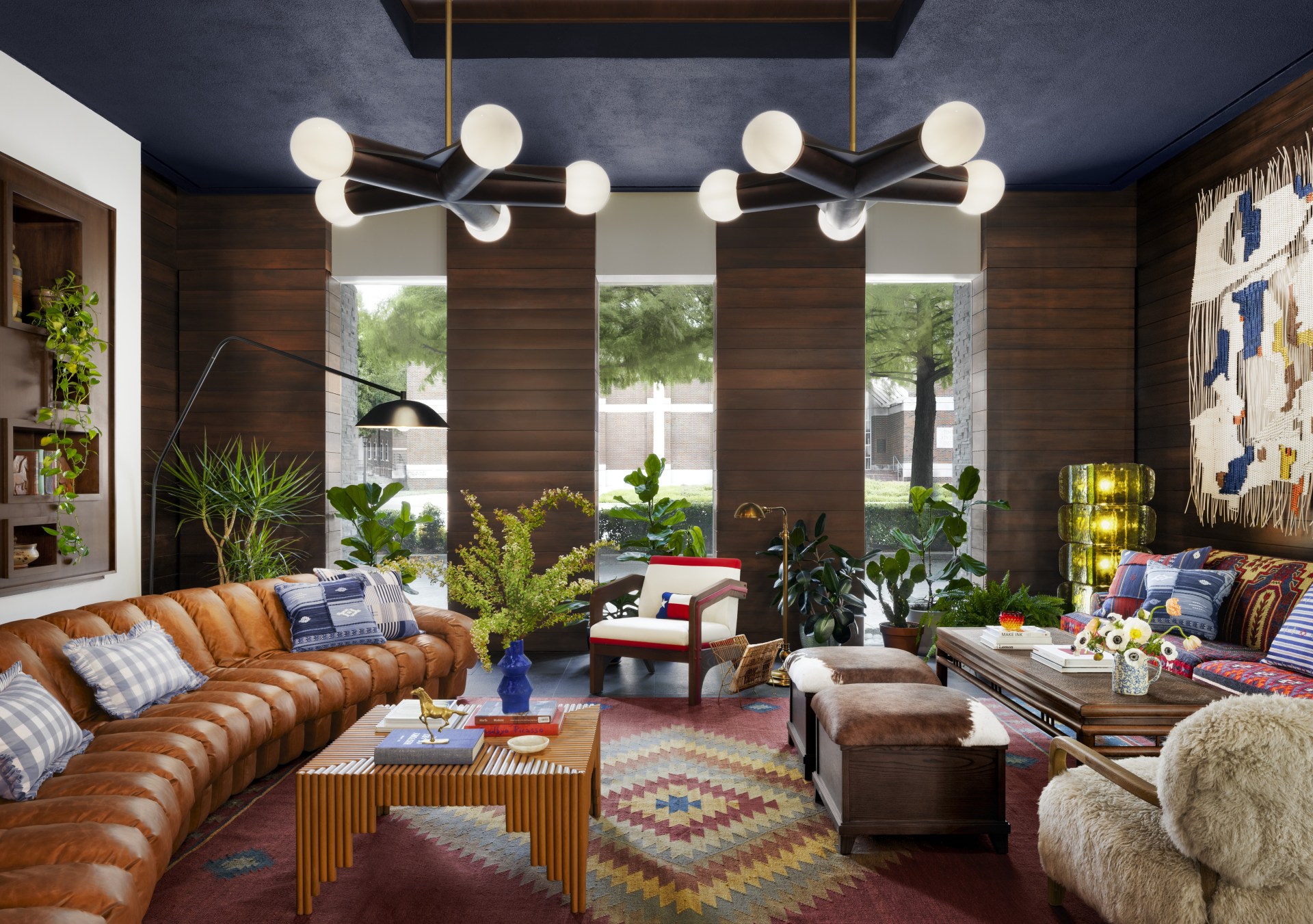 Graduate by Hilton Dallas, Lobby with accent chandelier light fixtures hanging from a dark colored ceiling in a room with wooden paneling and hanging artwork as well as recessed shelving on individual walls and lounge couches and accent tables and chairs with potted plants and floor lamps on an area rug, tanned leather and velvet sofas, cowhide ottomans, shearling armchairs, and handwoven textiles