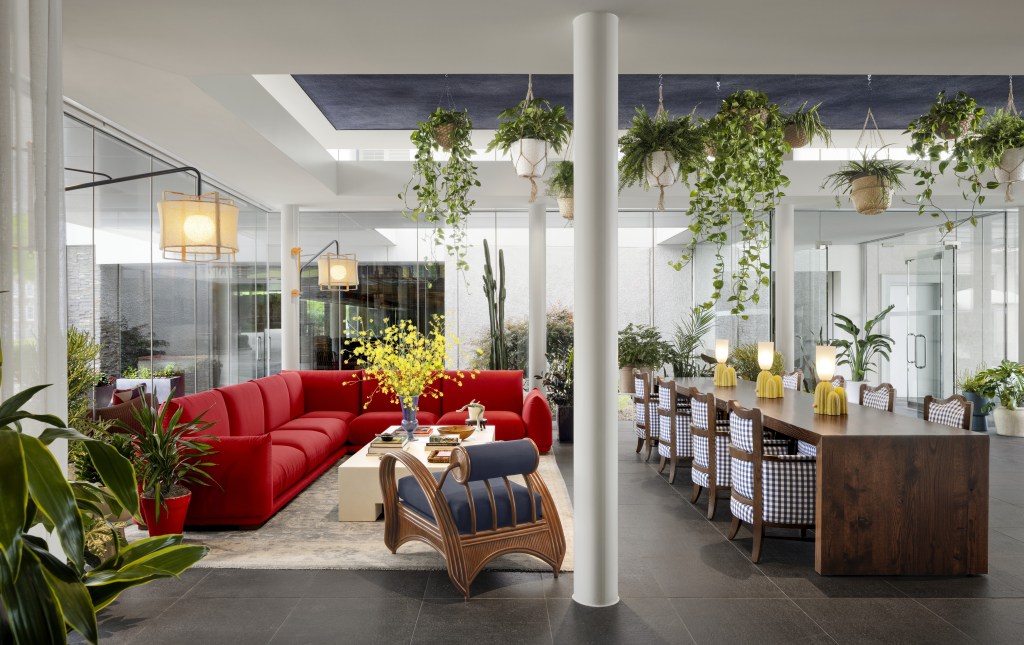 Graduate by Hilton Dallas, Lobby with a red couch a blue cushioned chair with wooden frame both on an area rug and an accent table with yellow flowers in a vase and floor lamps at each corner next to a long wooden table with yellow table lamps and chairs that have blue and white gingham print fabric with wooden frames and potted plants all around the room including some hanging from the ceiling