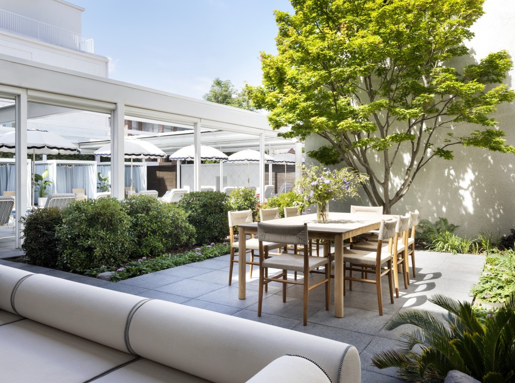 Graduate by Hilton Dallas, Outdoor Pool Area with lounge couch and dining table with chairs and a vase full of flowers on a patio surrounded by green bushes and trees and plants