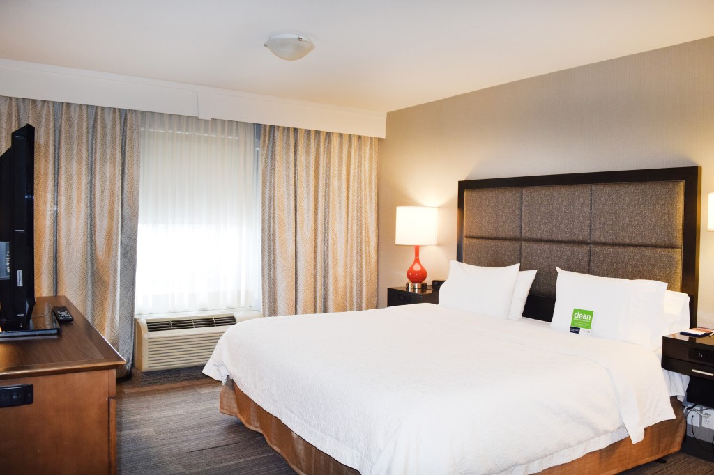Hampton Inn &amp; Suites by Hilton Calgary-University Northwest - King Suite Guest Room with white bed, television, large window, red table side lamp