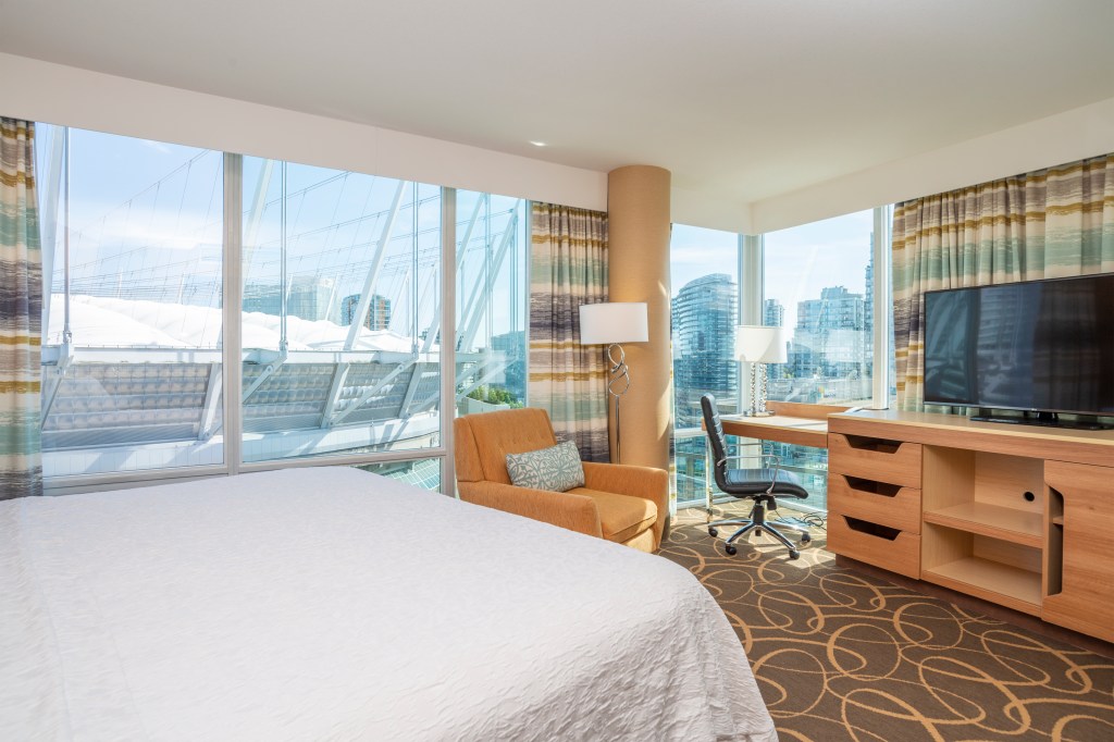Hampton Inn &amp; Suites by Hilton Vancouver-Downtown guest room with white bedding, large windows overlooking the city skyline, orange chair, desk and television