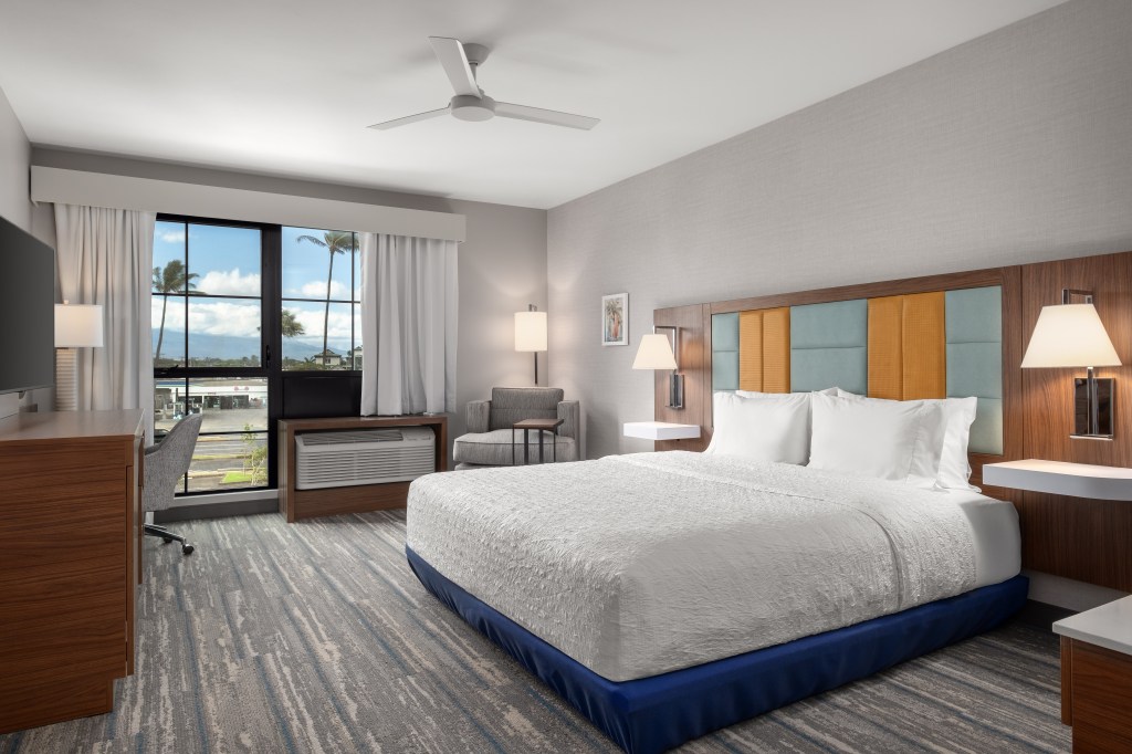 Hampton Inn and Suites Maui North Shore Guest Room with a bed facing a desk with chair next to a window and a dresser with a television mounted on the wall above across the room from a count with a table in front of it and artwork hanging on the wall above in a room with a ceiling fan