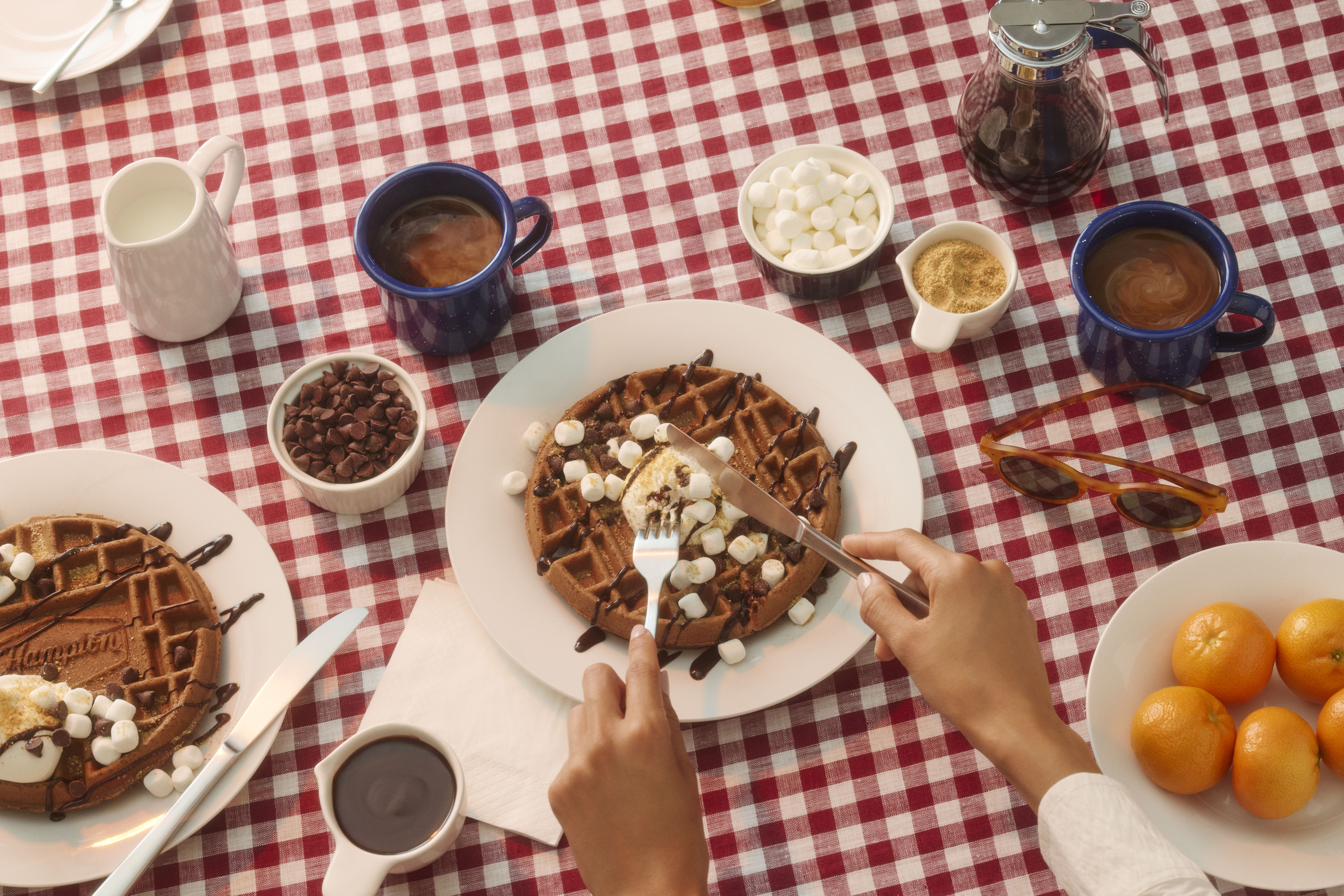Hampton by Hilton S’mores Waffles on a white and red checkered tablecloth, hands cutting a waffle with a knife and fork, waffles topped with chocolate and marshmallows, coffee and other breakfast items on the table