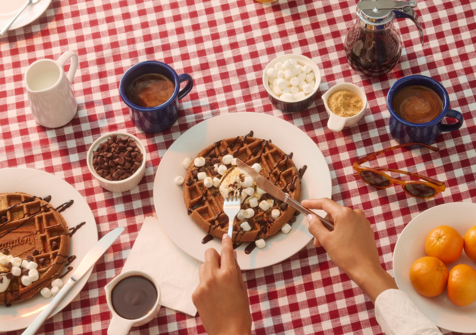 Hampton by Hilton S’mores Waffles on a white and red checkered tablecloth, hands cutting a waffle with a knife and fork, waffles topped with chocolate and marshmallows, coffee and other breakfast items on the table