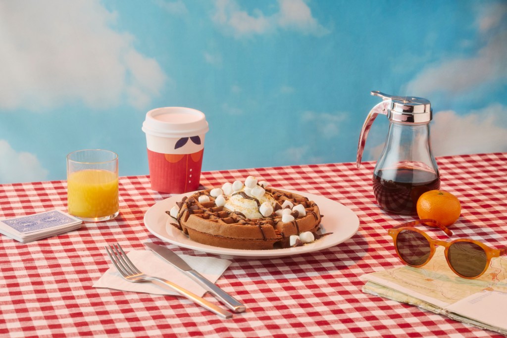 Hampton by Hilton S’mores Waffles on a red and white checkered table cloth with a knife and fork, napkin, coffee, orange juice, syrup, sunglasses, map, orange and deck of cards