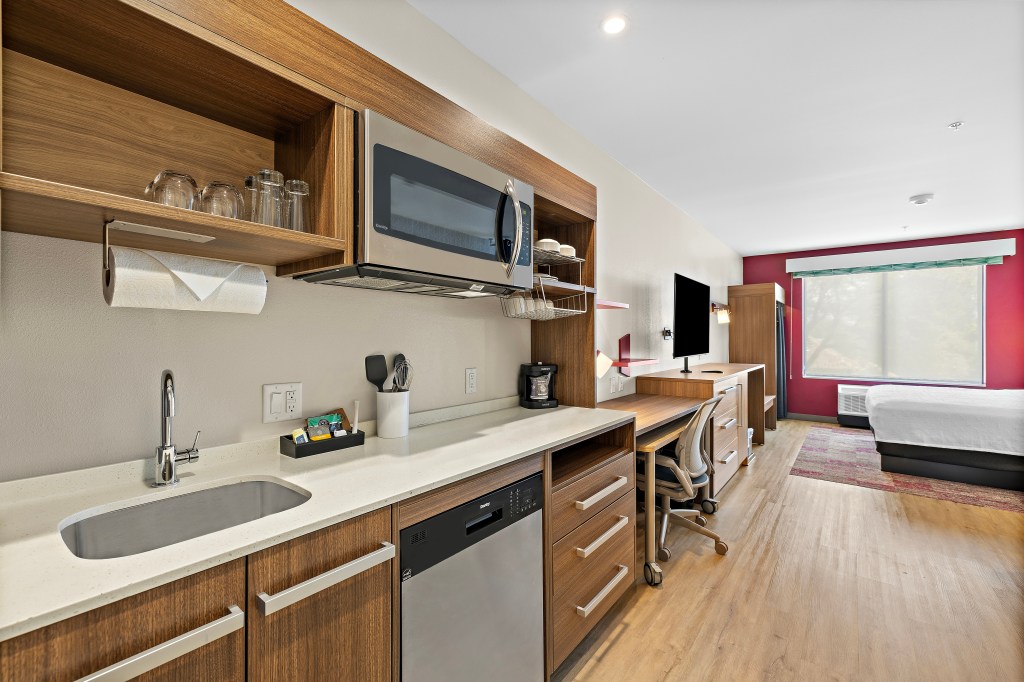 Home2 Suites by Hilton Riverside Downtown Guest Room Kitchen, white kitchen counter top, kitchen sink, dishwasher, under cabinet mounted paper towel holder, open shelving for kitchen dinnerware and glassware, microwave