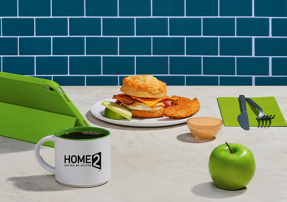 Home2 Suites by Hilton Build-Your-Own Breakfast Sandwiches - breakfast sandwich on a plate with apple slices and potatoes, table with green tablet, Home2 Suites by Hilton coffee mug, a green apple and cutlery