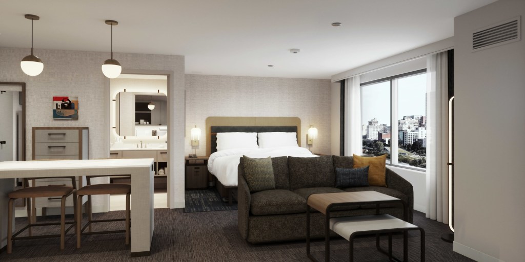 Homewood Suites by Hilton Fremont guest room with bed and end tables with mounted lighting above them next to a window and a couch sitting area with nested tables next to a bar table with two chairs and pendant lights above next to a dresser and a doorway to a bathroom with large mirror and vanity