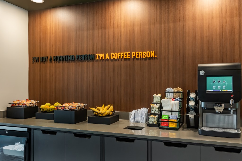 LivSmart Studios by Hilton Tullahoma - Breakfast Area with fruit, muffins, coffee and tea area and a words on the wall that read &quot;I'm not a morning person, I'm a coffee person&quot;