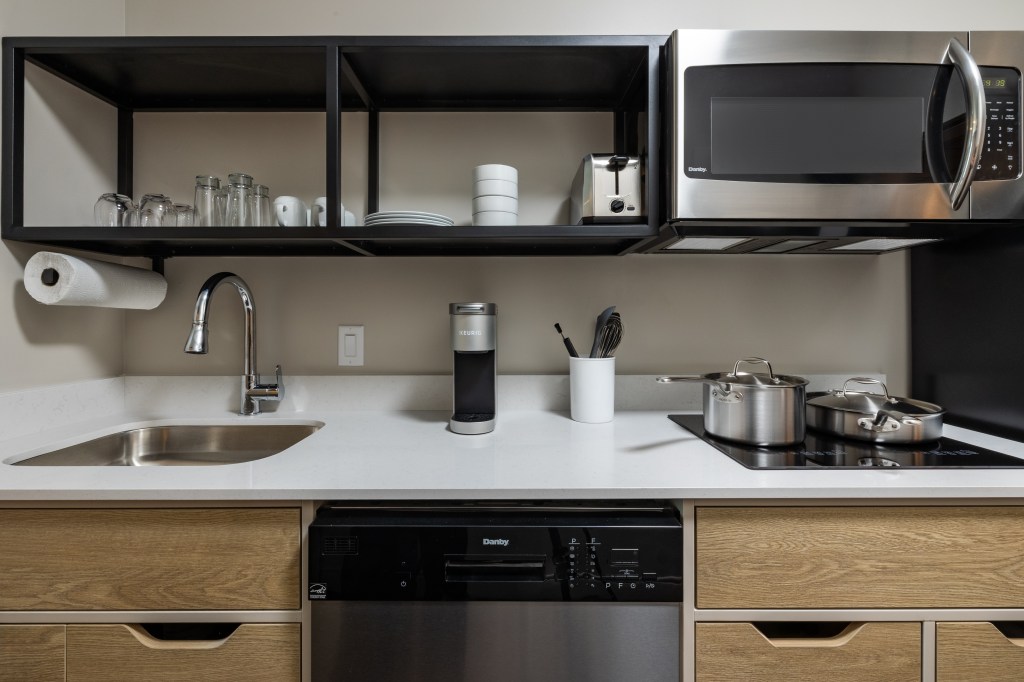LivSmart Studios by Hilton Tullahoma - Guest Room Kitchen with sink, dishwasher, stove top, microwave and open shelving with glasses, mugs, plates