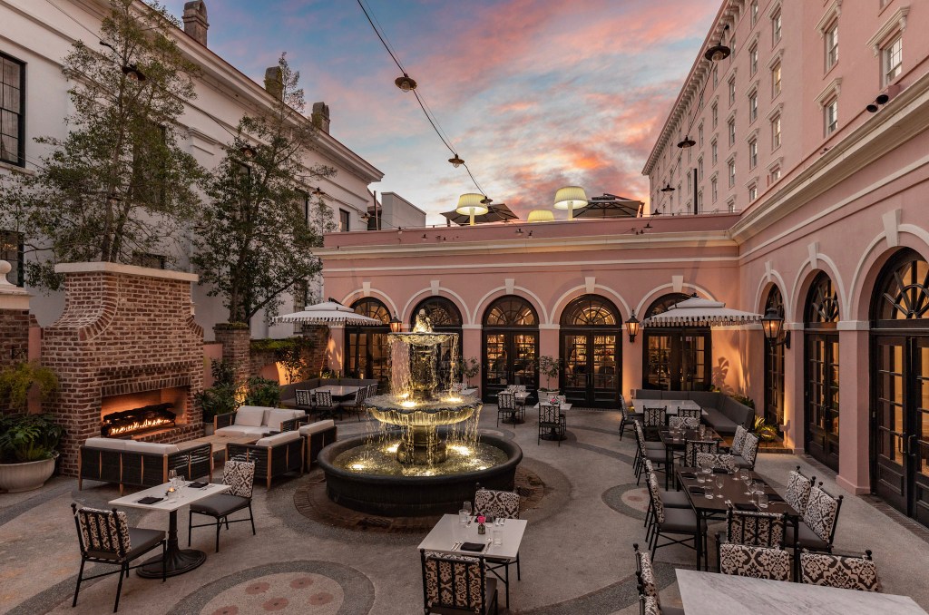 Romantic courtyard at Mills House Charleston with a central tiered fountain, brick fireplace, and pink historic architecture at dusk. Mills House Charleston, Curio Collection by Hilton