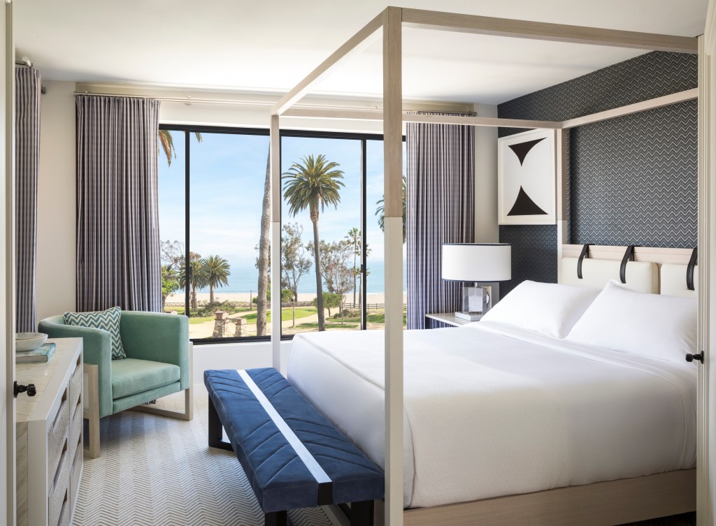 white bed, green chair and large windows overlooking palm trees, beach and ocean in Signature Oceanfront Suite at Oceana Santa Monica, LXR Hotels & Resorts