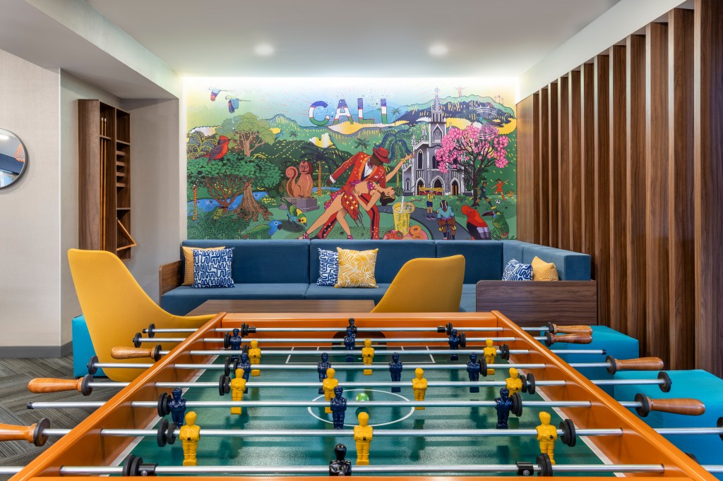 Tru by Hilton Cali Sur lounge area with a foosball table with a are mural on the wall behind a blue couch with yellow and blue printed throw pillows and yellow chairs