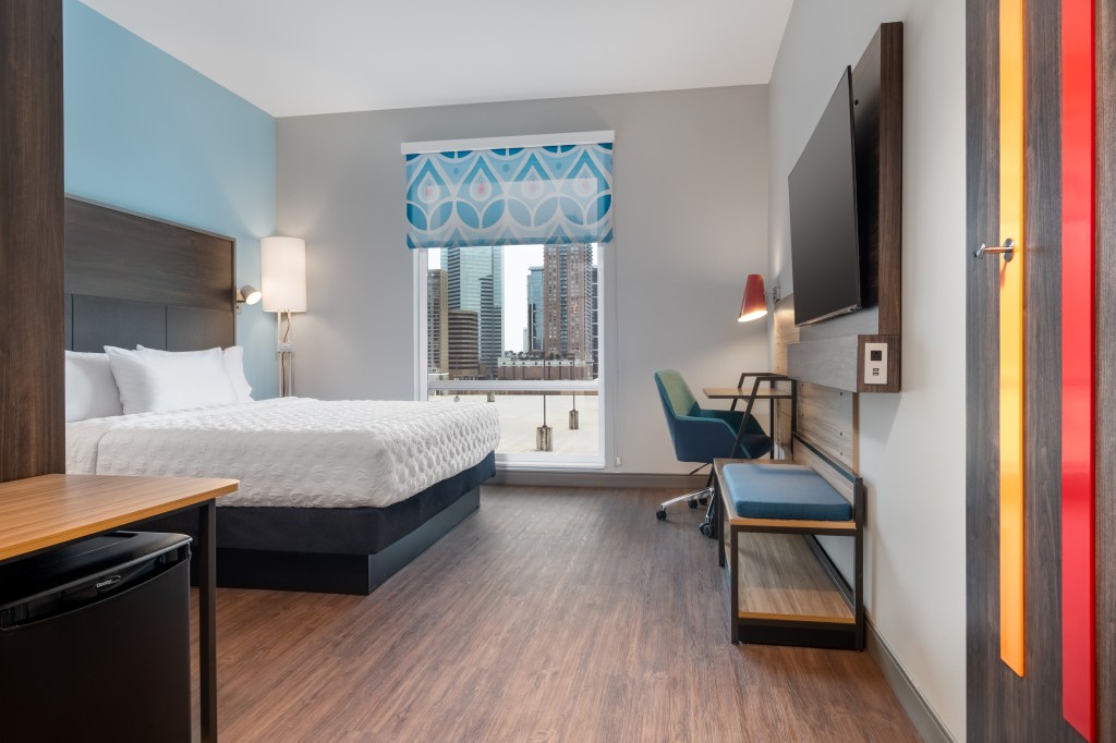 Tru by Hilton Houston Downtown Convention Center guest room with a bed with lamps and a desk with a chair and a mounted television and mini fridge