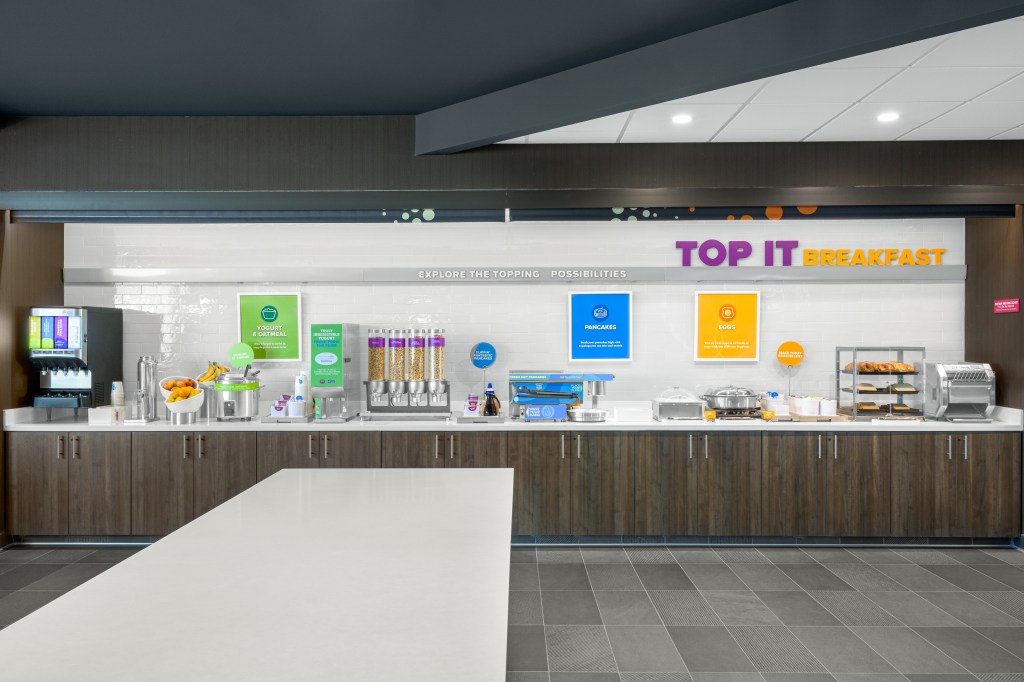 Tru by Hilton Norco Eastvale breakfast bar with a beverage fountain machine next to cups and bowls with fresh fruit in them including bananas and oranges next to a crock pot with a ladle for serving and cereal dispenser containers next to a pancake making machine with a syrup container next to the machine and warming containers and a clear case with pastries on display next to a toaster
