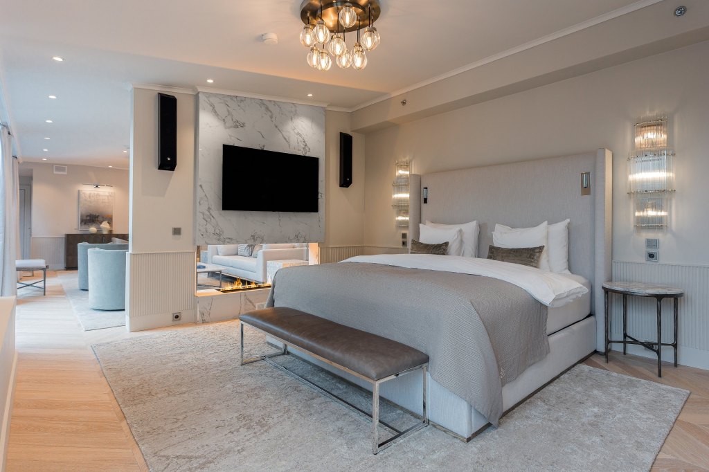 Waldorf Astoria Helsinki - Penthouse Suite Bedroom, silver, marbel and brown leather decor,tv and speaker system over fireplace, bed with bench at the foot, side tables and sconces, view of living room in background