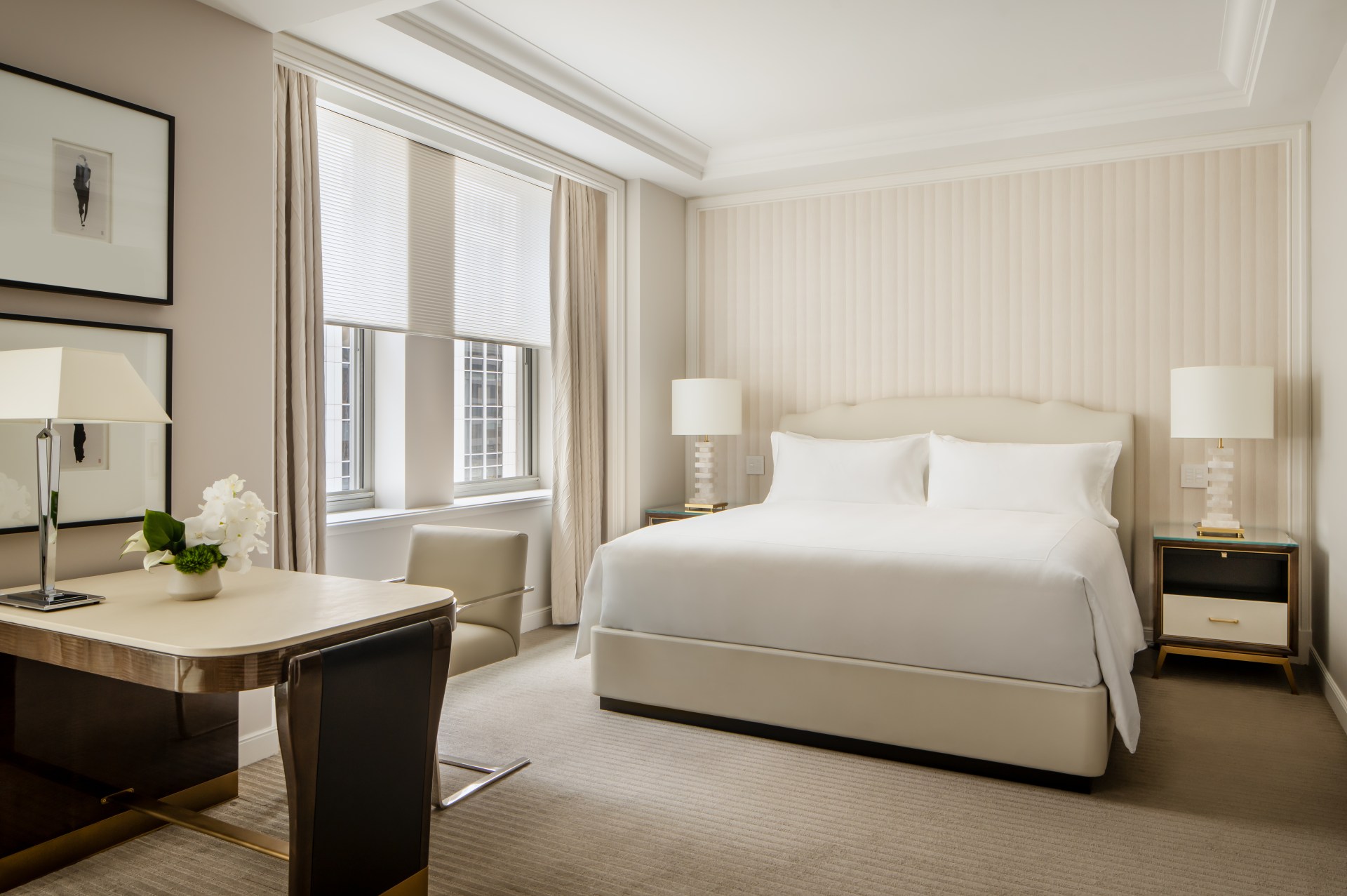 Waldorf Astoria New York Park Avenue Junior Suite Bed room with framed artwork on a wall next to a window by the bed with white bedding and two nightstands with table lamps on them and a desk chair and desk with a lamp and flowers in a vase