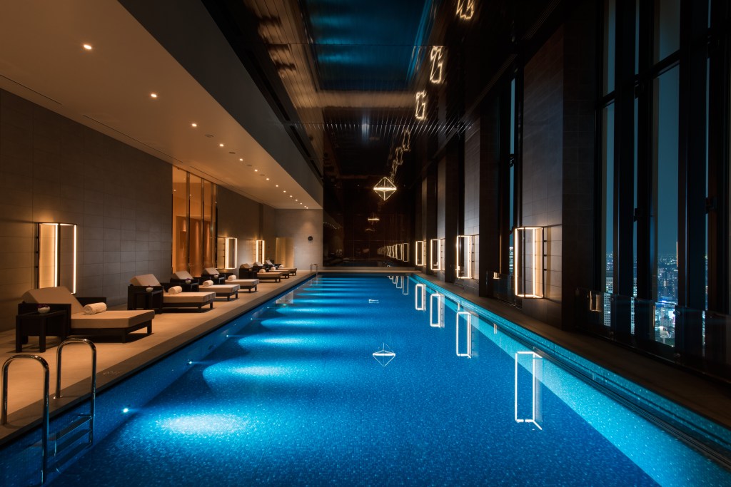 Conrad Osaka indoor pool with dim lighting throughout and chaise pool chairs facing the pool with floor to ceiling windows overlooking the city, Indoor Pool, blue tiled floor, chaise seating, rolled white towels, sconce wall light fixtures, city view