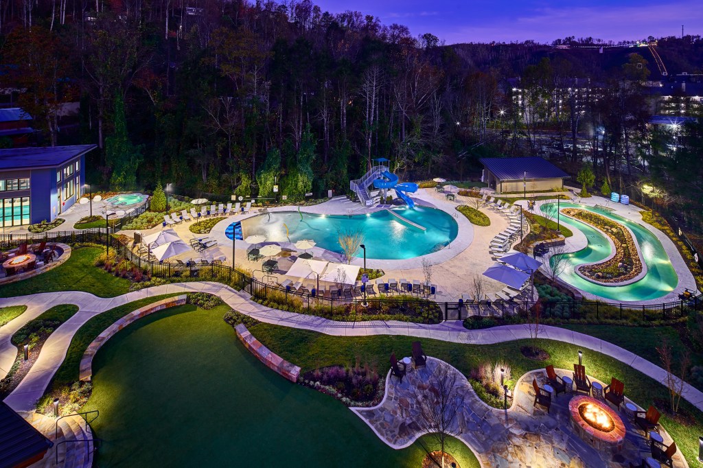 Embassy Suites by Hilton Gatlinburg Resort Pool at Night with a firepit surrounded by red adironack chairs separated by a fence in the pool area are three separate pools one off shaped circle lazy river float pool next to a pool with lounge pool chairs and umbrellas with a blue water slide next to a smaller plunge hot tub size pool not far from another firepit area