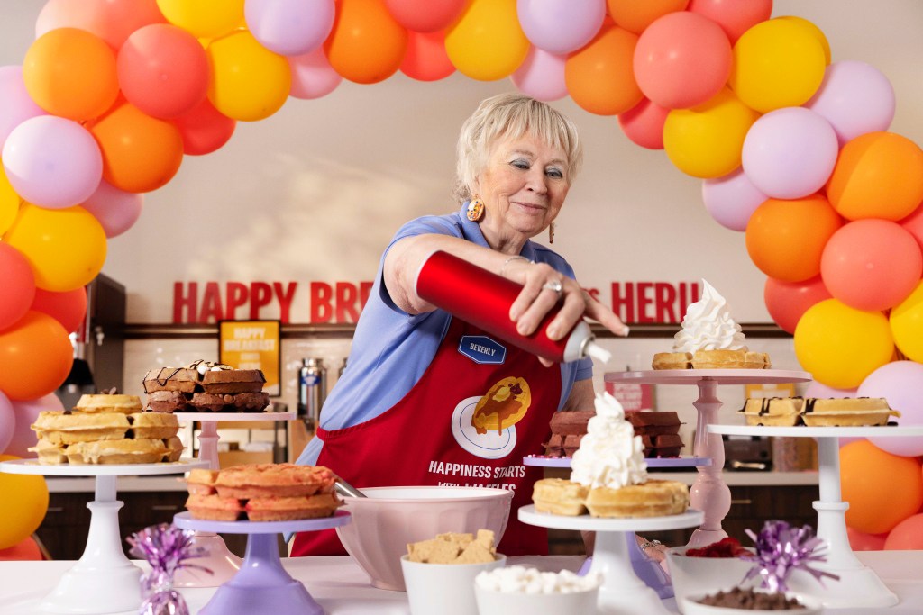 Hampton by Hilton Waffle Boss Beverly Christmas adds whipped cream to a waffle while surrounded by waffles, waffle toppings and colorful balloons