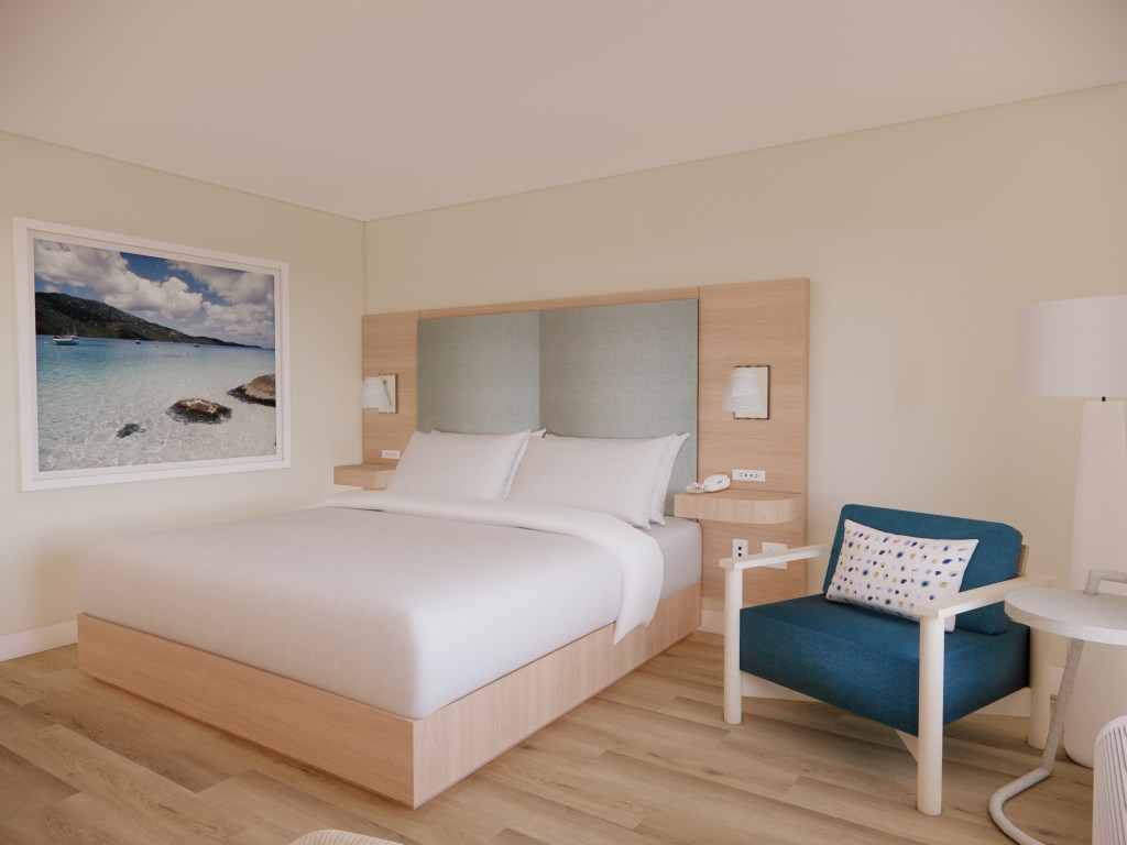 Hampton by Hilton St. Thomas Guest Room with photo hanging on wall in a white frame next to bed with floating wooden shelves and mounted white lighting next to a blue cushioned chair with a table and lamp