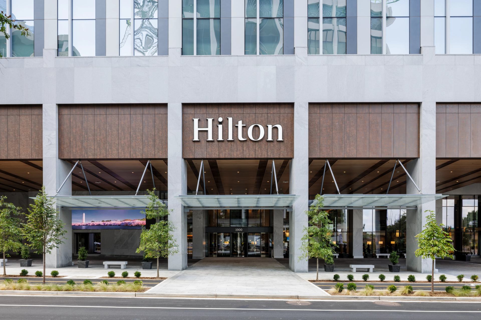 Hilton Arlington Rosslyn The Key Exterior Porte-cochère entrance with large sidewalk and pull through for drop off with trees and potted plants next to bench seating