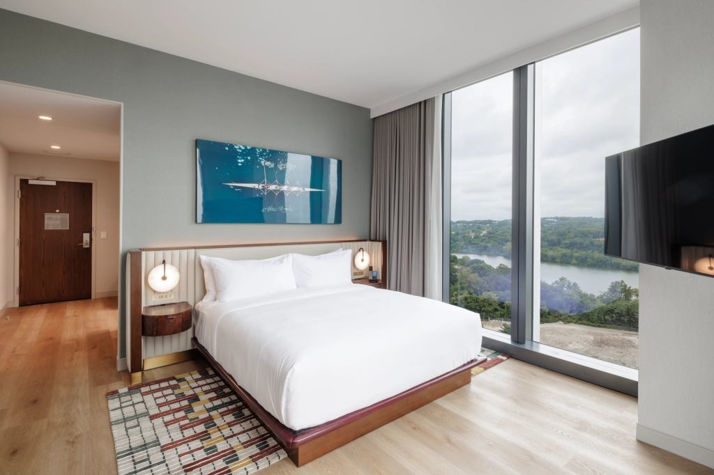 Hilton Arlington Rosslyn The Key King Guest Room with floor to ceiling window next to bed with a white upholstered headboard with round mounted lighting and wooden floating end tables with a gold baseboard with white bedding and a green red yellow and neutral colored area rug under it and a picture of crew boat hanging on wall above the bed